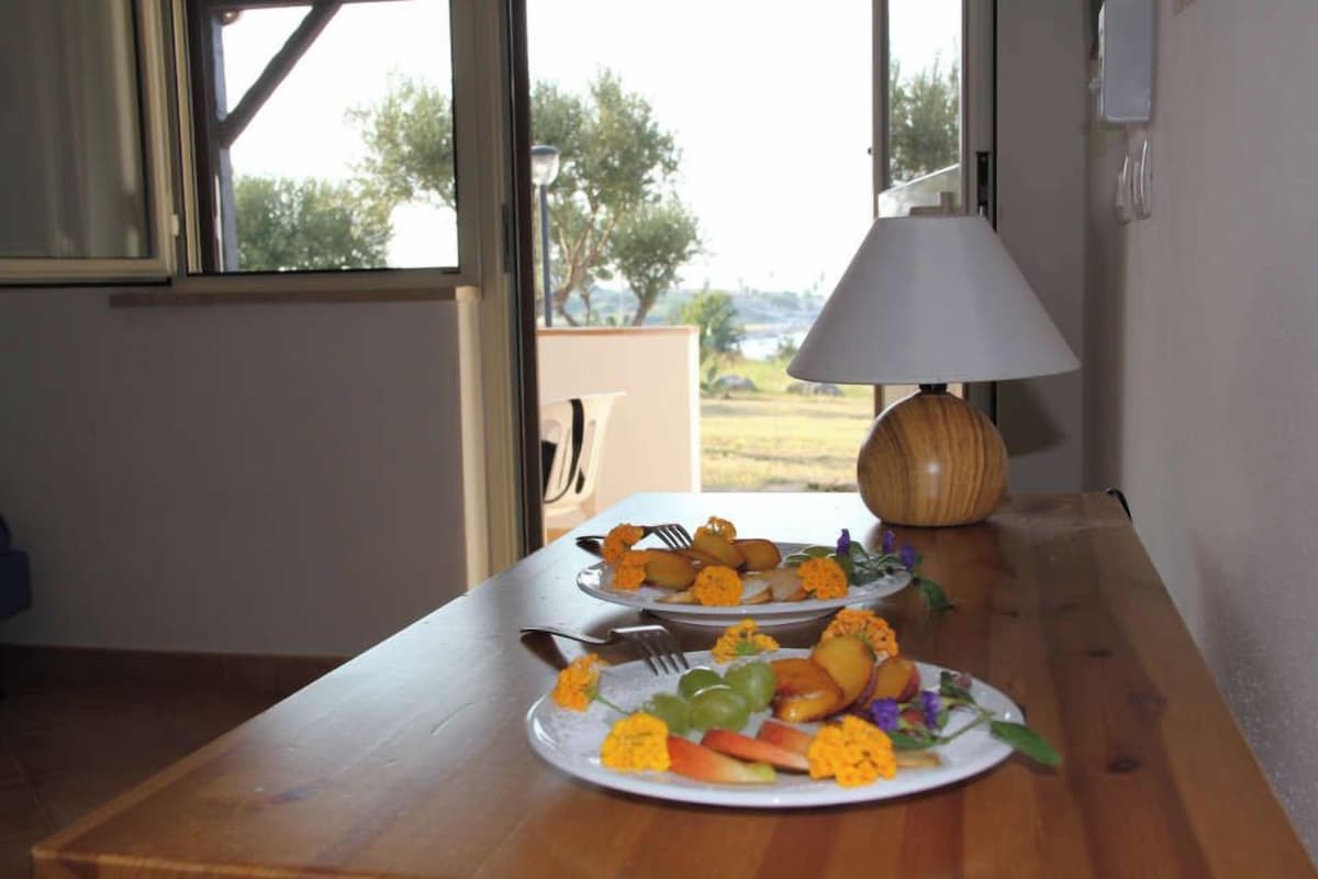 A dining table with plates of fruit and flowers, a lamp, and a view of trees through a window.