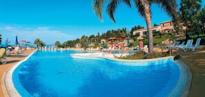 A large swimming pool with lounge chairs and umbrellas, surrounded by palm trees and a building in the background. - Villaggio Dolomiti sul Mare (Briatico)