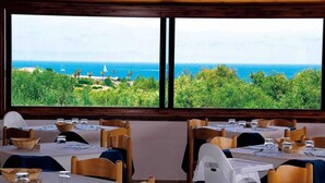 Un restaurant dont les tables sont dressées pour le service, avec de grandes fenêtres offrant une vue sur l’océan et la verdure.