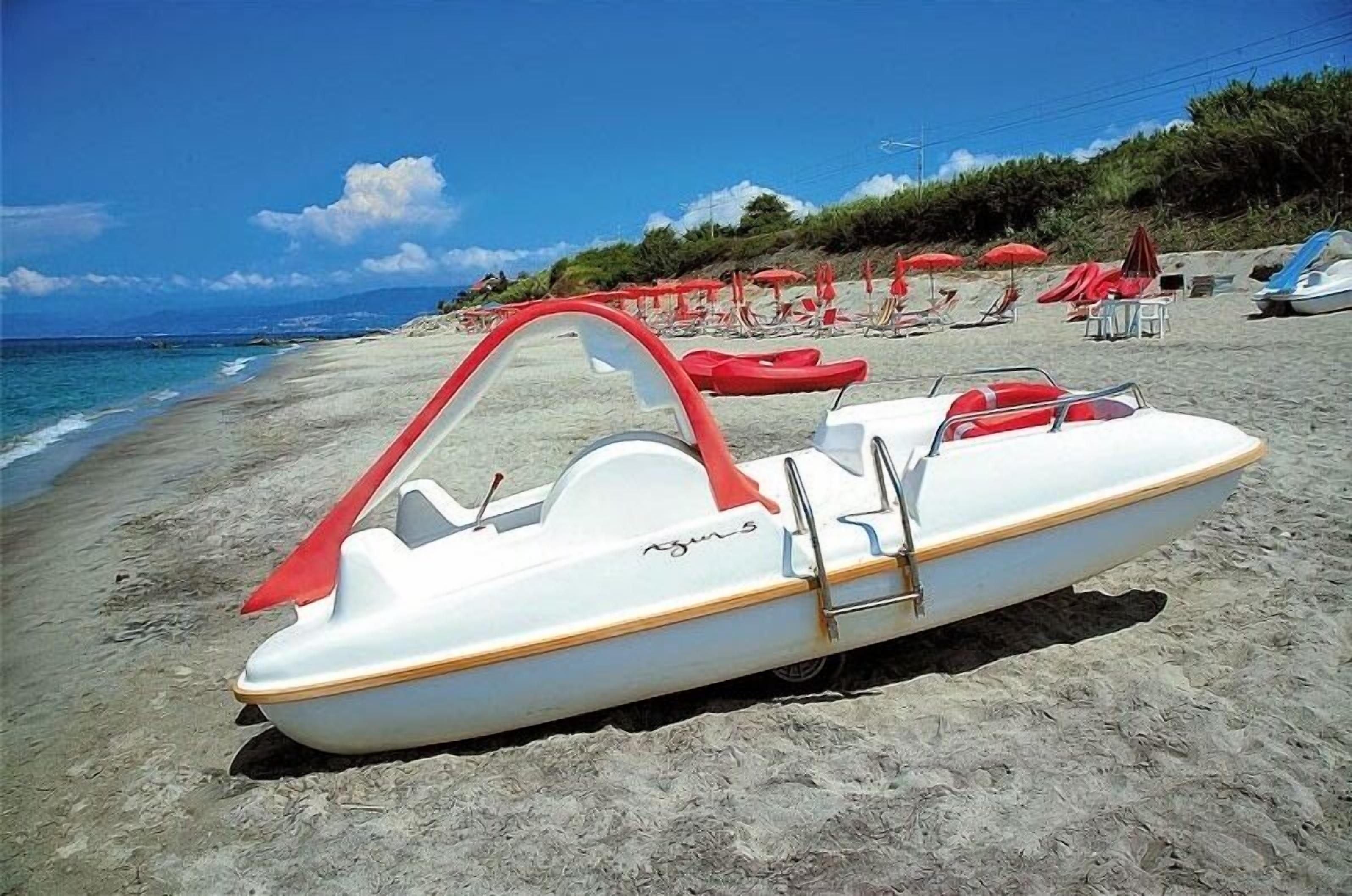 A small white boat with red accents on a sandy beach, surrounded by red umbrellas and beach chairs.