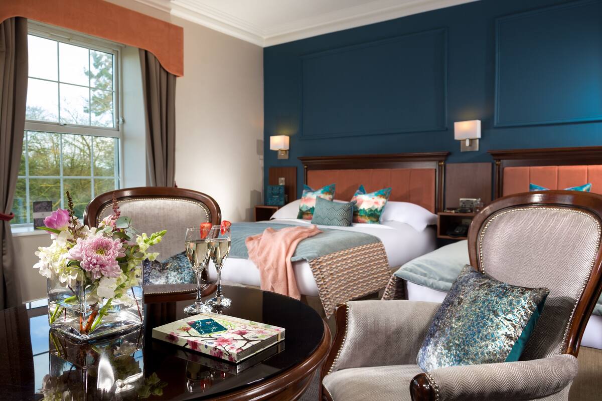 Una habitación de hotel con una cama, dos sillas, una mesa con un jarrón de flores y un libro.