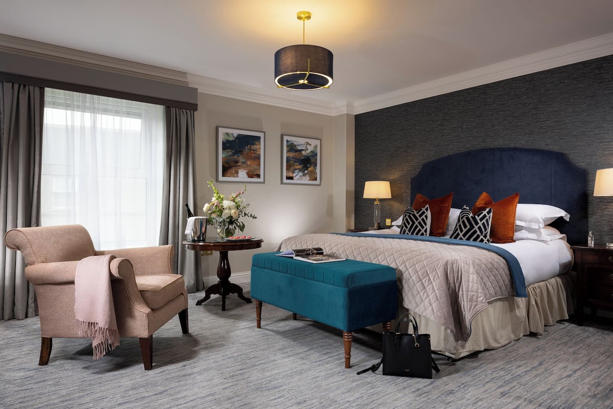 Habitación de hotel con una cama grande, una silla, una mesita de noche con un florero y vistas al exterior.