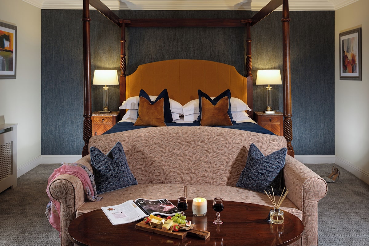 Una cama con dosel, con un edredón estampado y almohadas, una mesita de noche con lámpara, una pequeña mesa con una revista, velas y aperitivos.
