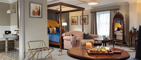 Habitación de hotel con cama con dosel, una zona de estar con sofá y sillones, una mesa de comedor con fruta y copas de vino, y un espejo grande.