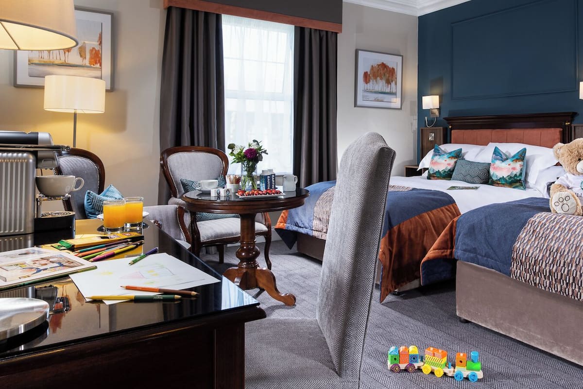 Habitación de hotel con dos camas, un escritorio, una silla, una lámpara y un osito de peluche sobre una de las camas.