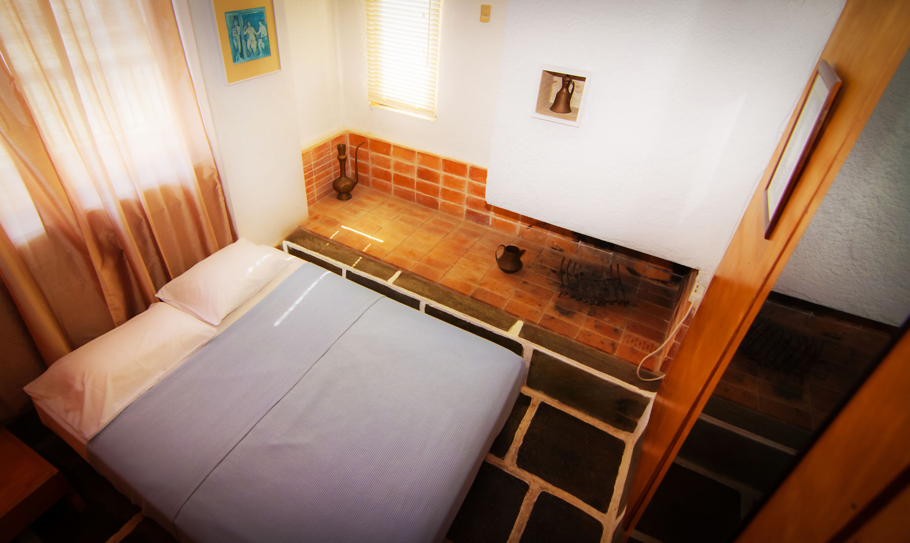 A small, compact room with a bed, a small table, and a window with blinds.