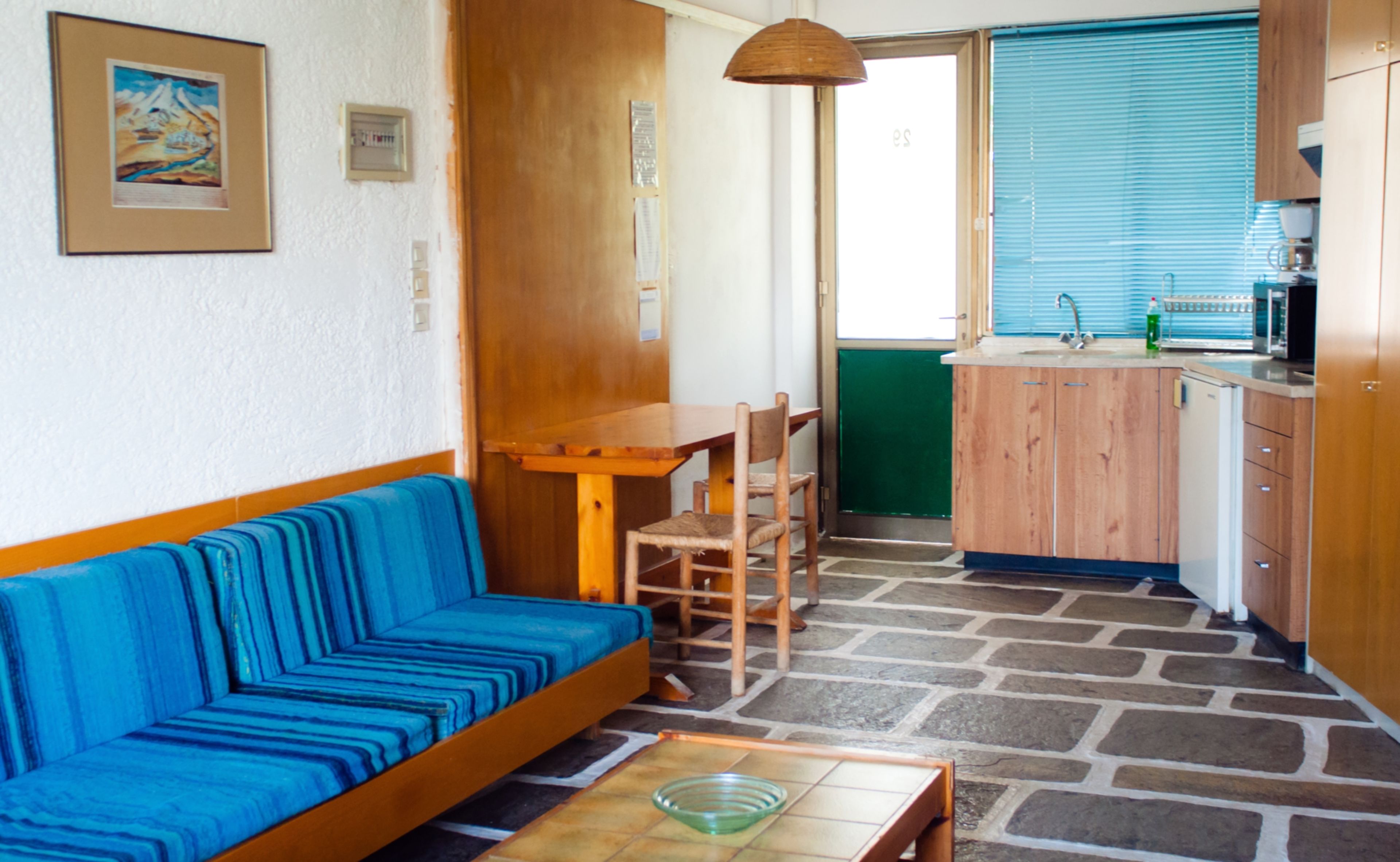 Une pièce avec un canapé bleu, une table en bois, un coin cuisine doté de meubles en bois et un sol en pierre.