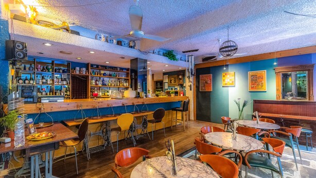 A bar area with wooden tables and chairs, shelves with bottles, and a ceiling fan. - Apollonia Hotel Apartments (Vari-Voula-Vouliagmeni)