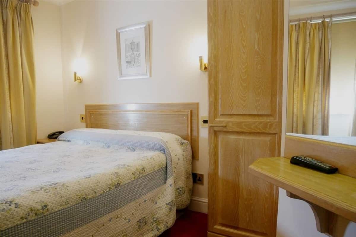 Habitación de hotel con cabecera de madera, cama con ropa de cama floral, mesita de noche con un control remoto y un espejo sobre la mesa.