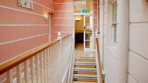 Un couloir avec un escalier en bois, un panneau indiquant la sortie de secours et une vue sur une pièce dotée d’une fenêtre.