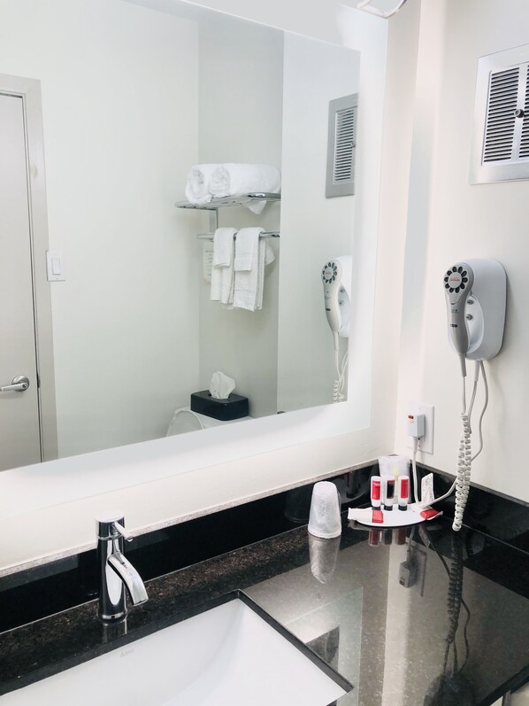 A hotel bathroom with a large mirror, a sink, a towel rack with towels, a hairdryer, and toiletries on the counter.