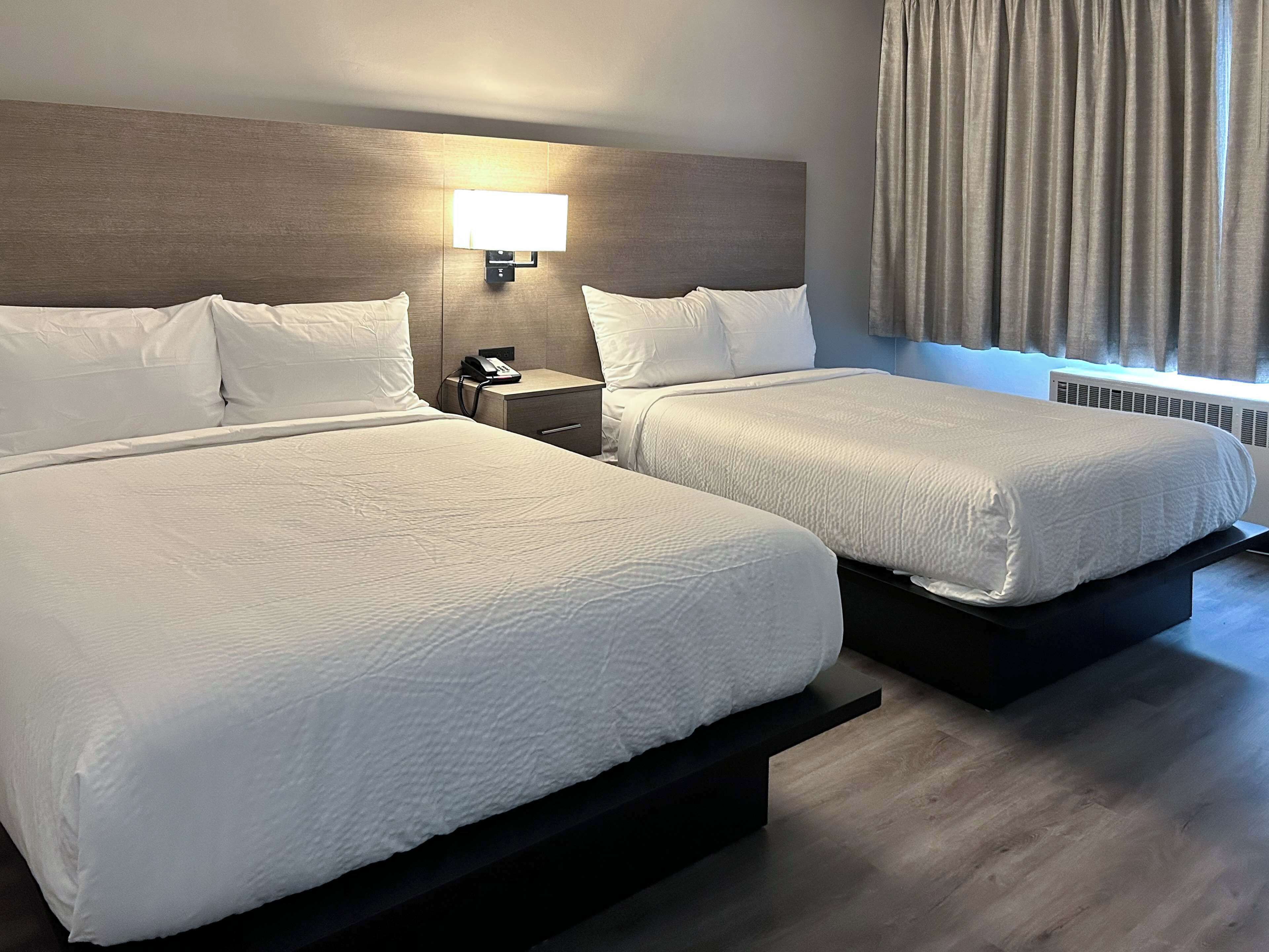 A hotel room with two beds, a nightstand with a phone, a wall-mounted lamp, and a window with curtains.