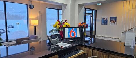 A reception desk with a computer, phone, and a vase of flowers. There is a window with a view of a parking lot and a building outside.