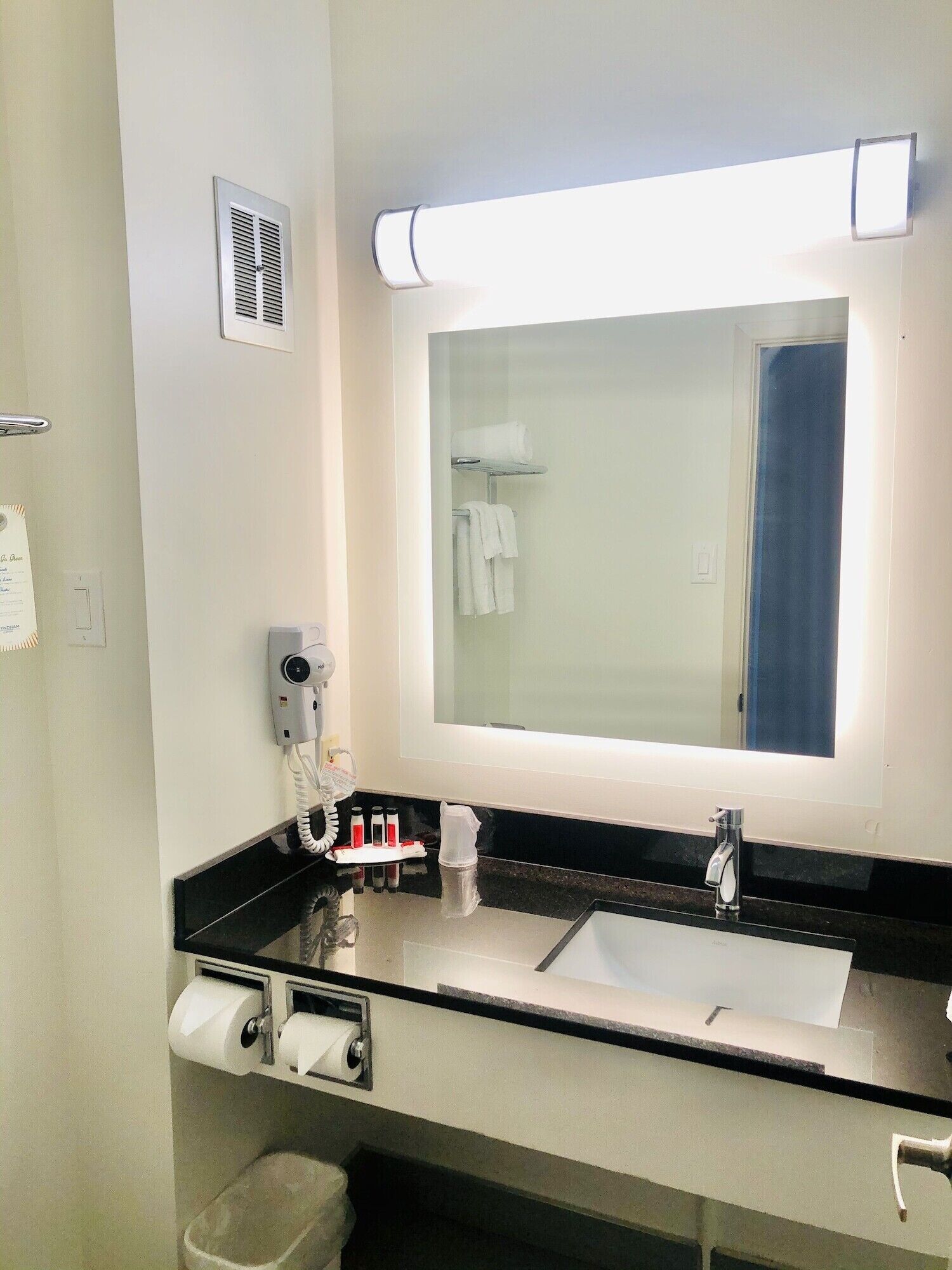 A hotel bathroom with a large mirror, a sink, a towel rack, a hand dryer, and a trash bin.