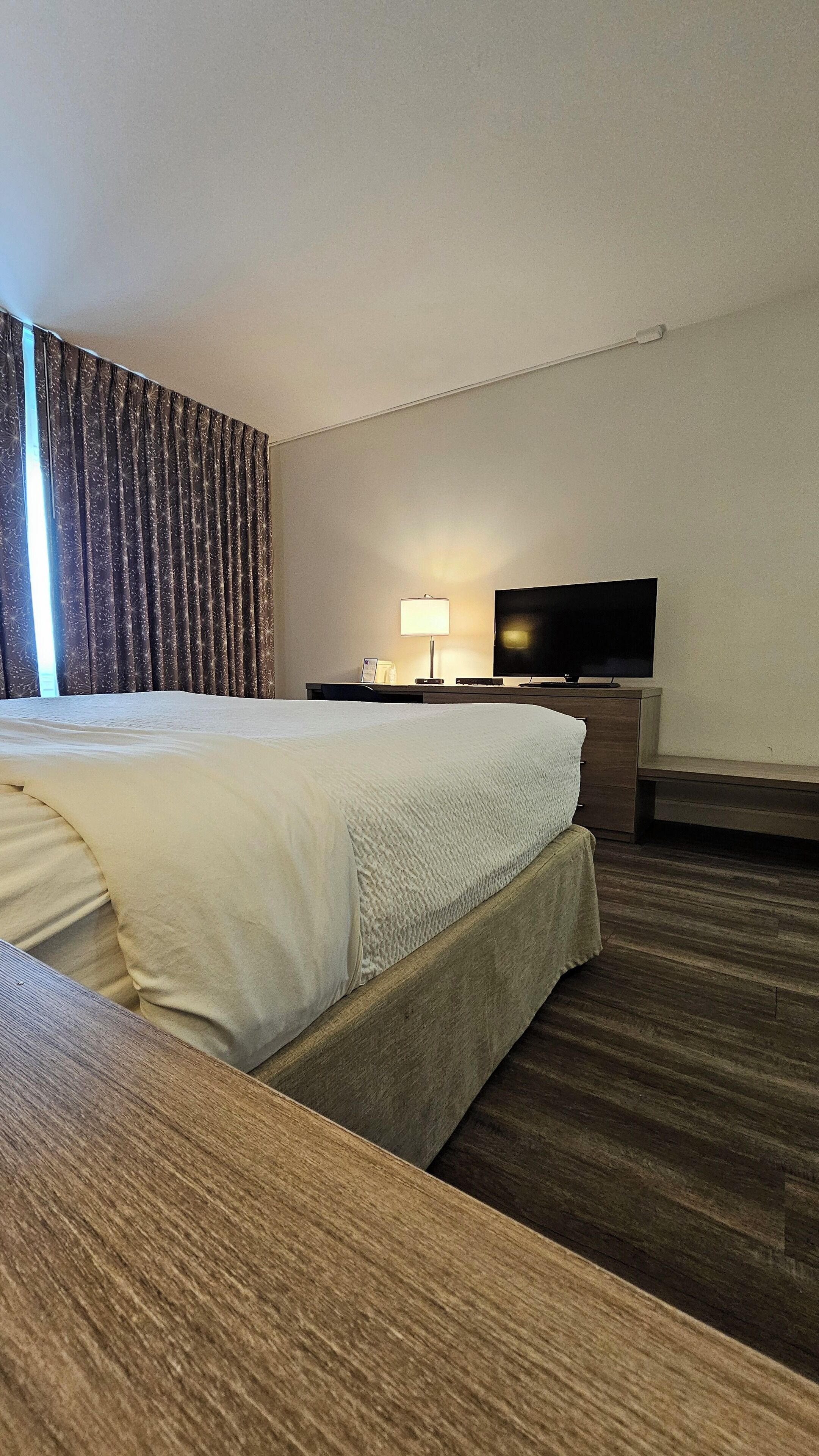 A hotel room with a large bed, a flat-screen TV, a bedside table with a lamp, and a wooden bench.