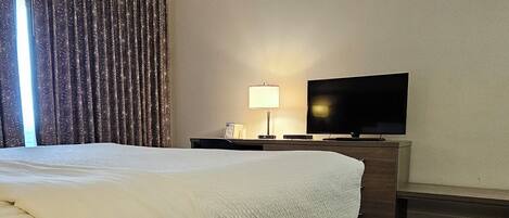 A hotel room with a large bed, a flat-screen TV, a bedside table with a lamp, and a wooden bench.