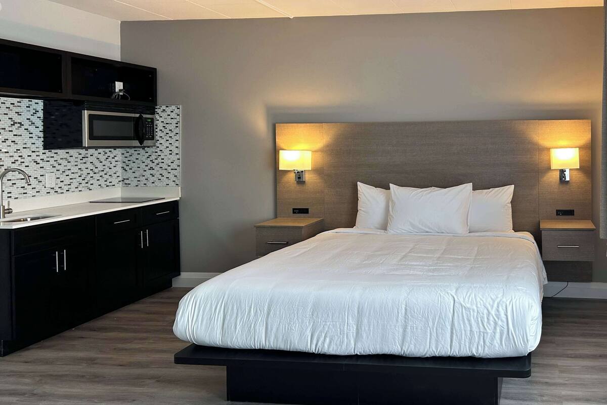 Une chambre d’hôtel moderne dotée d’un grand lit, de tables de chevet avec lampes, d’un four à micro-ondes encastré et d’un coin cuisine avec un dosseret carrelé.