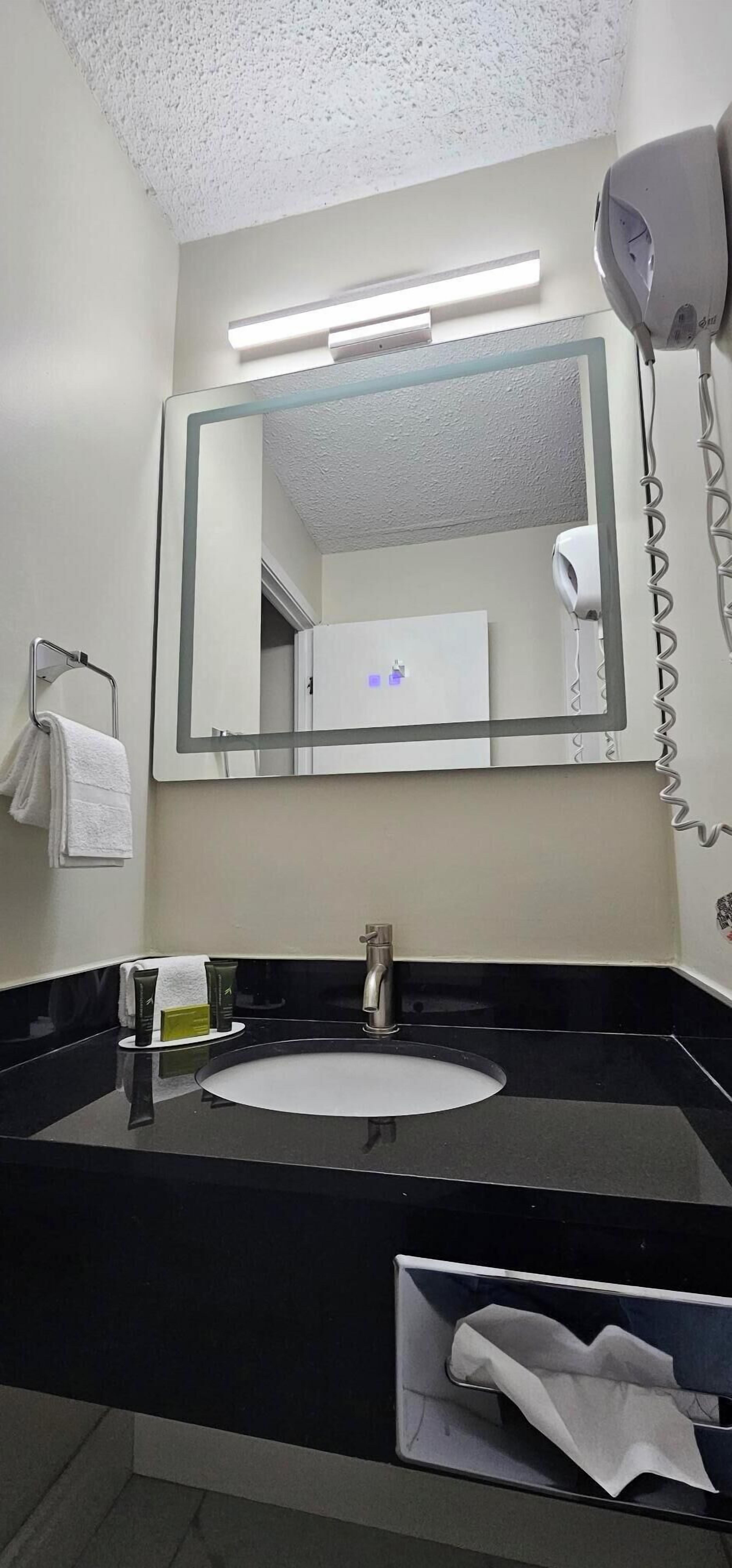 Un baño moderno con encimera negra, un lavabo redondo, un espejo grande, un secador de pelo y un perchero para toallas.