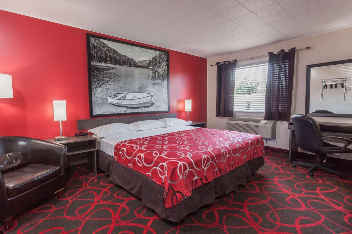 A hotel room with a bed, a desk, a chair, a large window with curtains, and a painting on the wall.