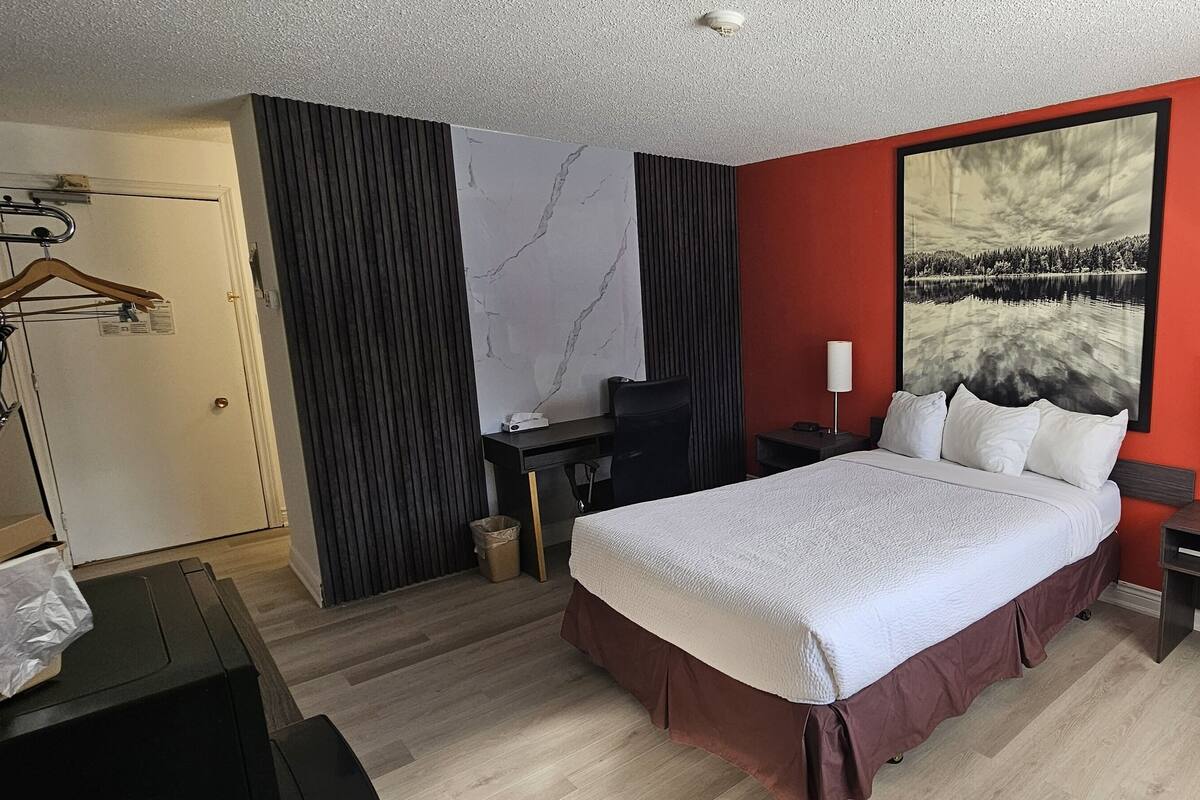 A hotel room with a bed, bedside tables, a desk, a chair, a wardrobe, and a large painting on the wall.