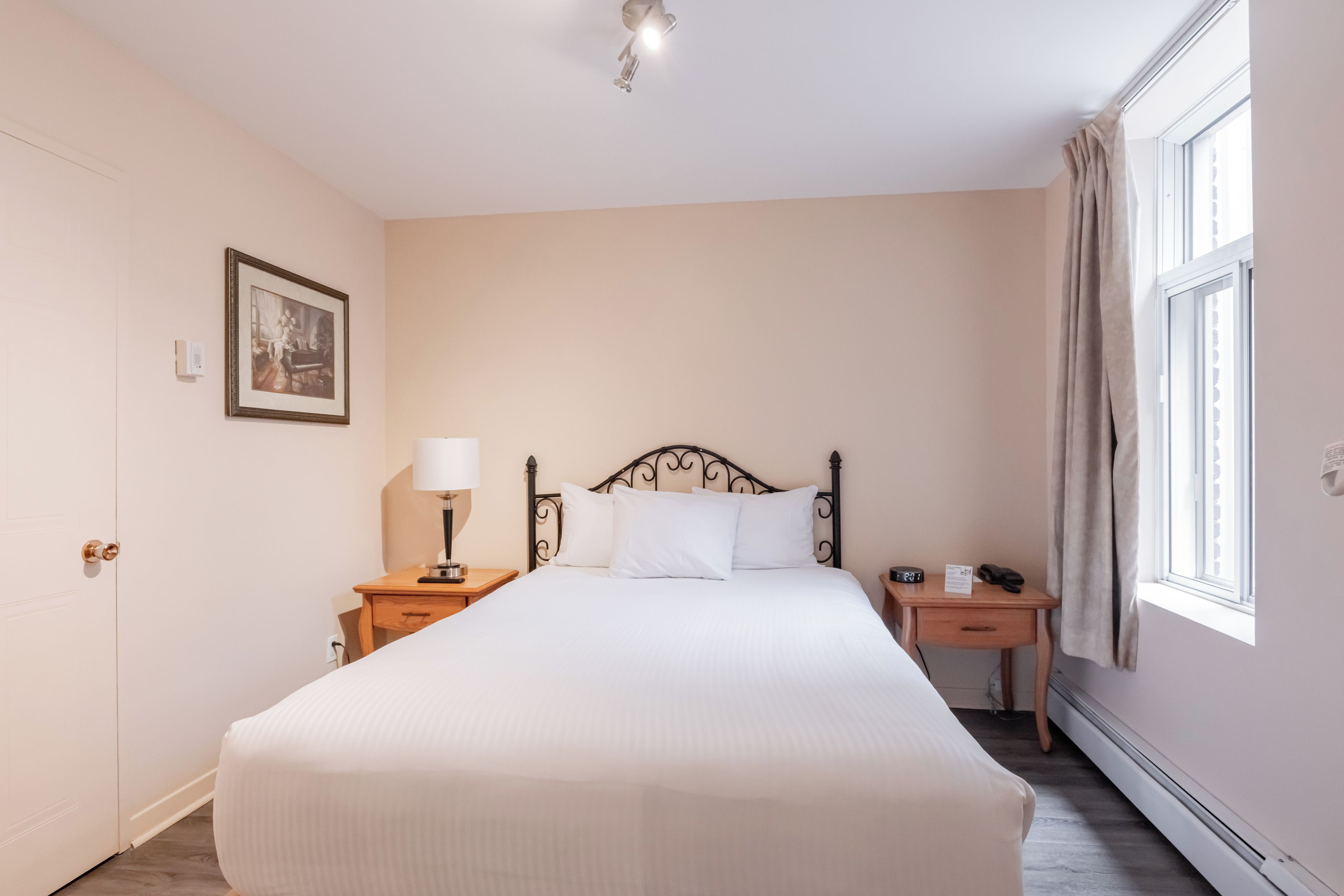Una habitación de hotel con una cama grande, una mesita de noche con lámpara, un tocador con reloj, un cuadro en la pared y una ventana con cortinas.