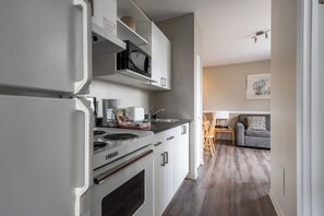 A modern kitchen with white cabinets, a built-in microwave, and a stove. There is a dining area with a gray sofa and a table. - Hotel Le Roberval (Montreal)
