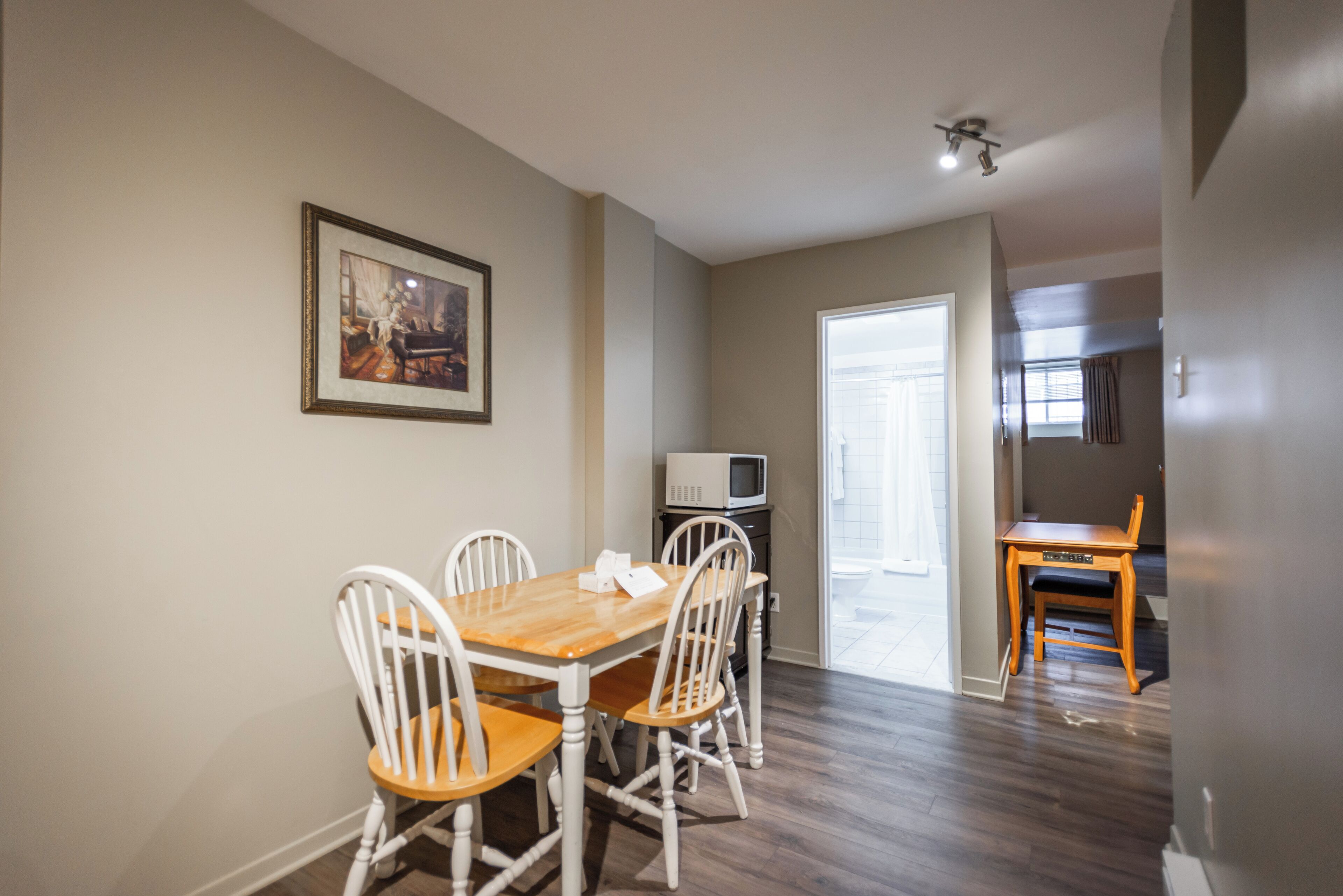 A dining area with a wooden table and white chairs, a framed picture on the wall, and a microwave on a cabinet.