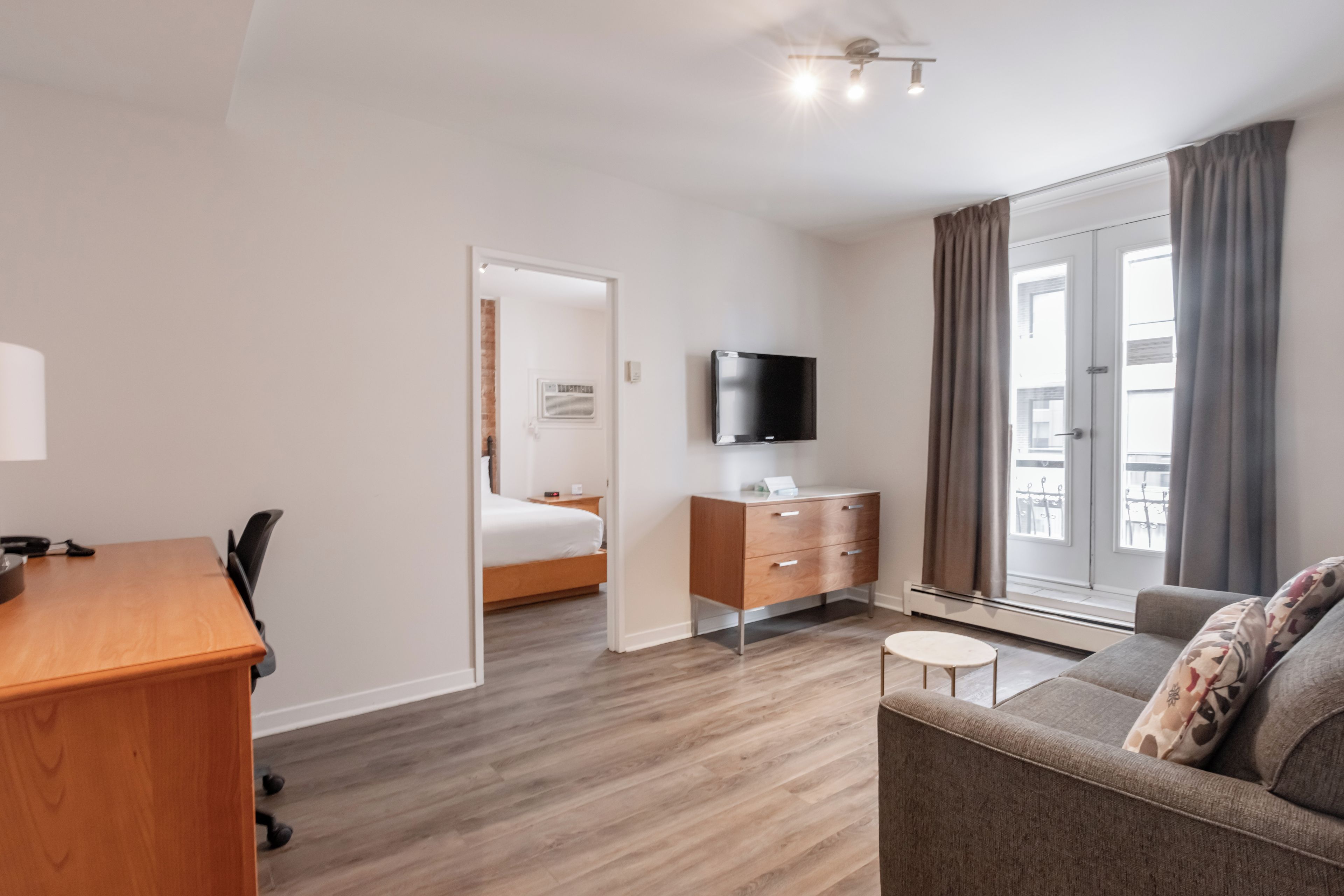 A hotel room with a wooden desk, a bed, a TV, a sofa, and a window with curtains.