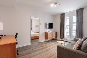 A hotel room with a wooden desk, a bed, a TV, a sofa, and a window with curtains. - Hotel Le Roberval (Montreal)