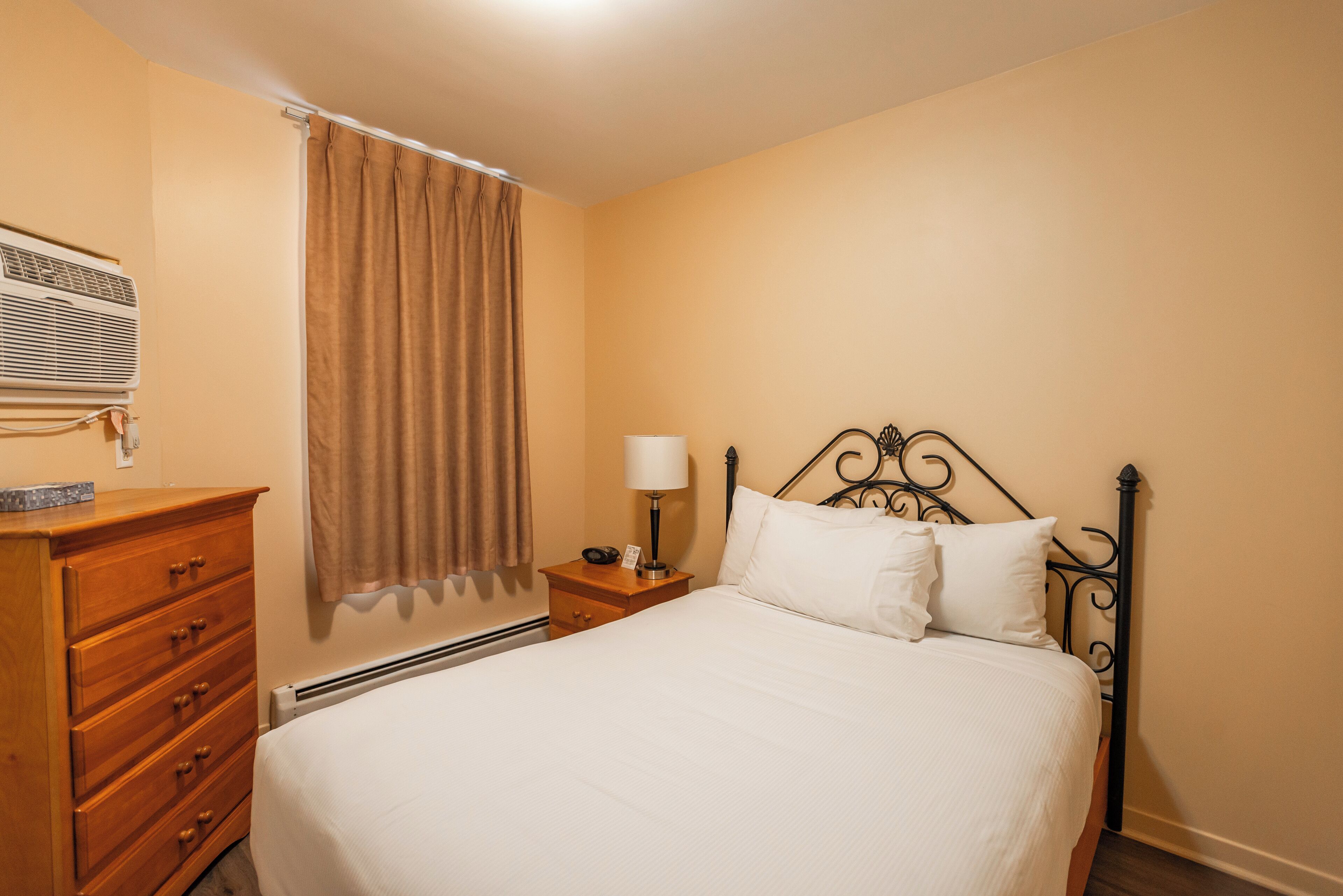 A hotel room with a bed, a dresser, a nightstand with a lamp, and a window with curtains.