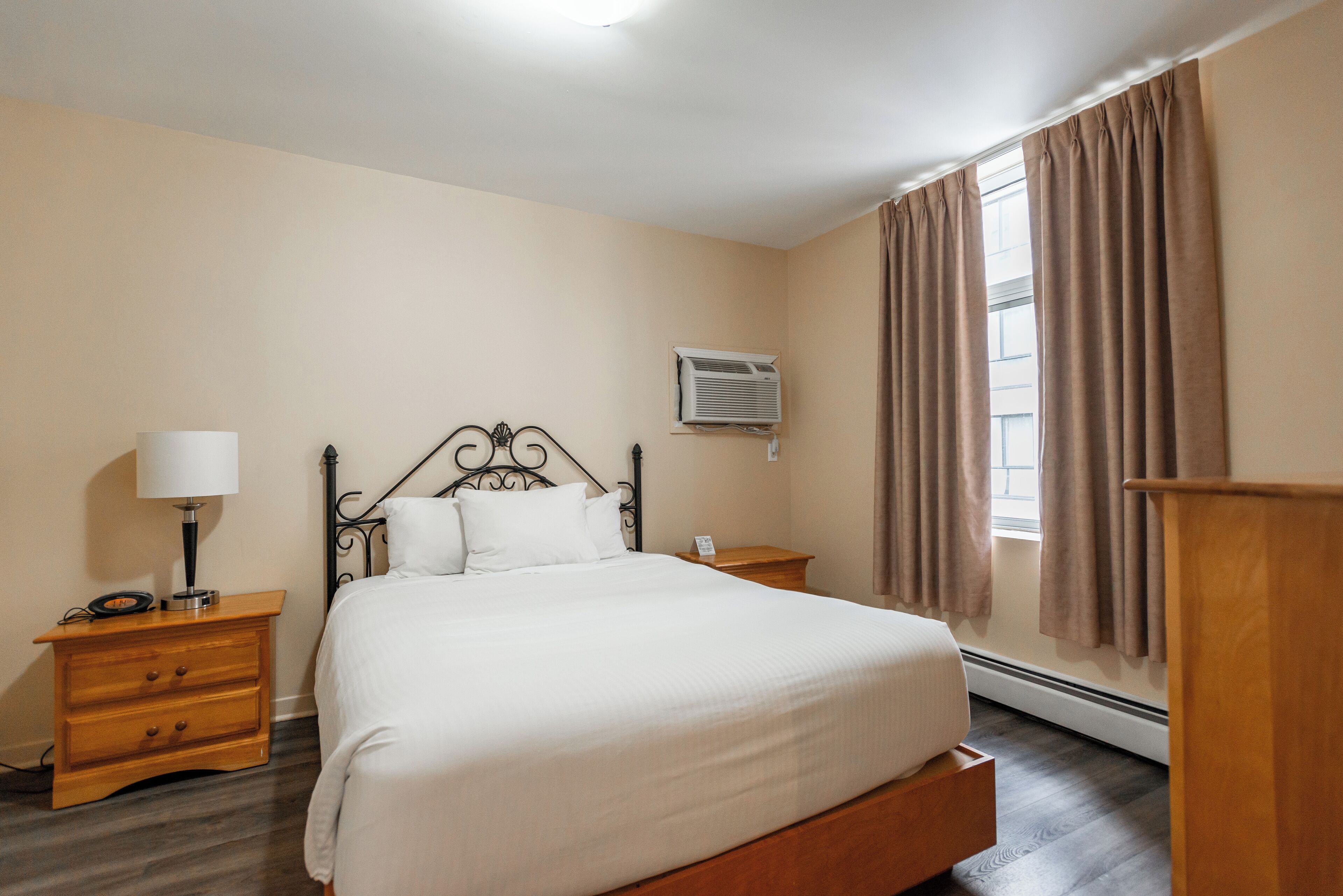 Une chambre à coucher bien rangée, avec un lit, des tables de chevet, une lampe, un appareil de climatisation et une fenêtre avec des rideaux.