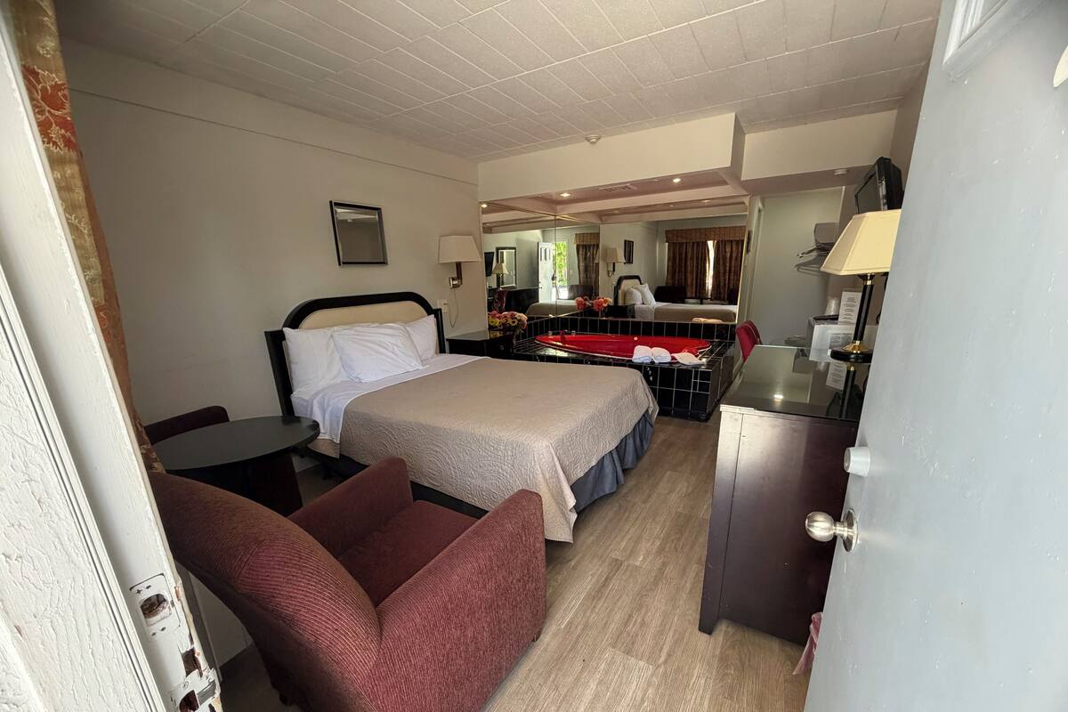 A hotel room with a bed, a desk, a chair, a small table, a mirror, and a bathtub.
