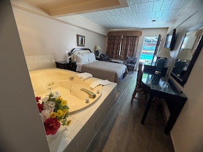 A hotel room with a large bathtub, a bed, a desk with a chair, a television, and a window with curtains.