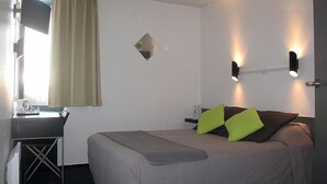 Habitación de hotel con cama, escritorio y silla. Se aprecia una ventana con cortinas, un espejo montado en la pared y dos apliques.