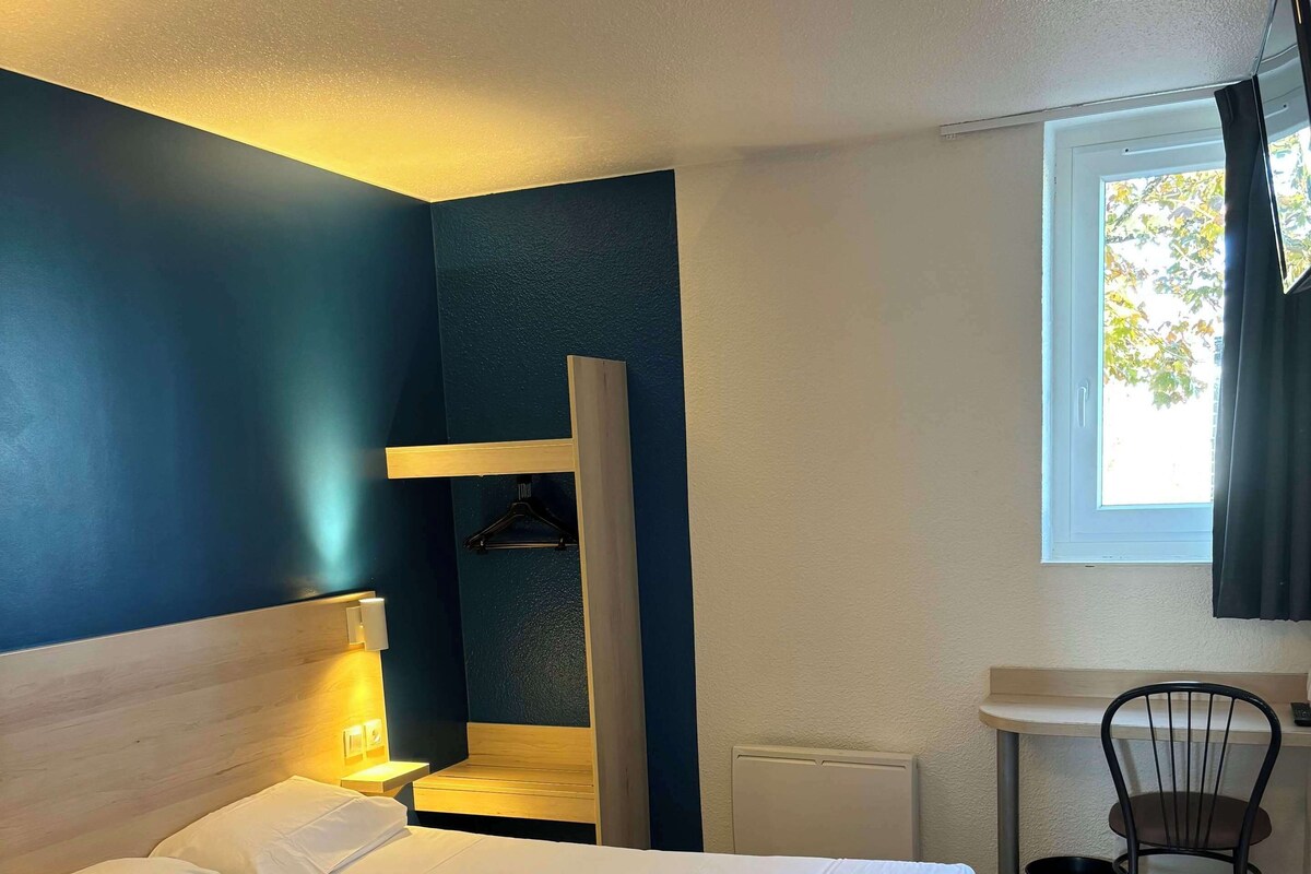 A hotel room with a bed, a small table, a chair, and a window.