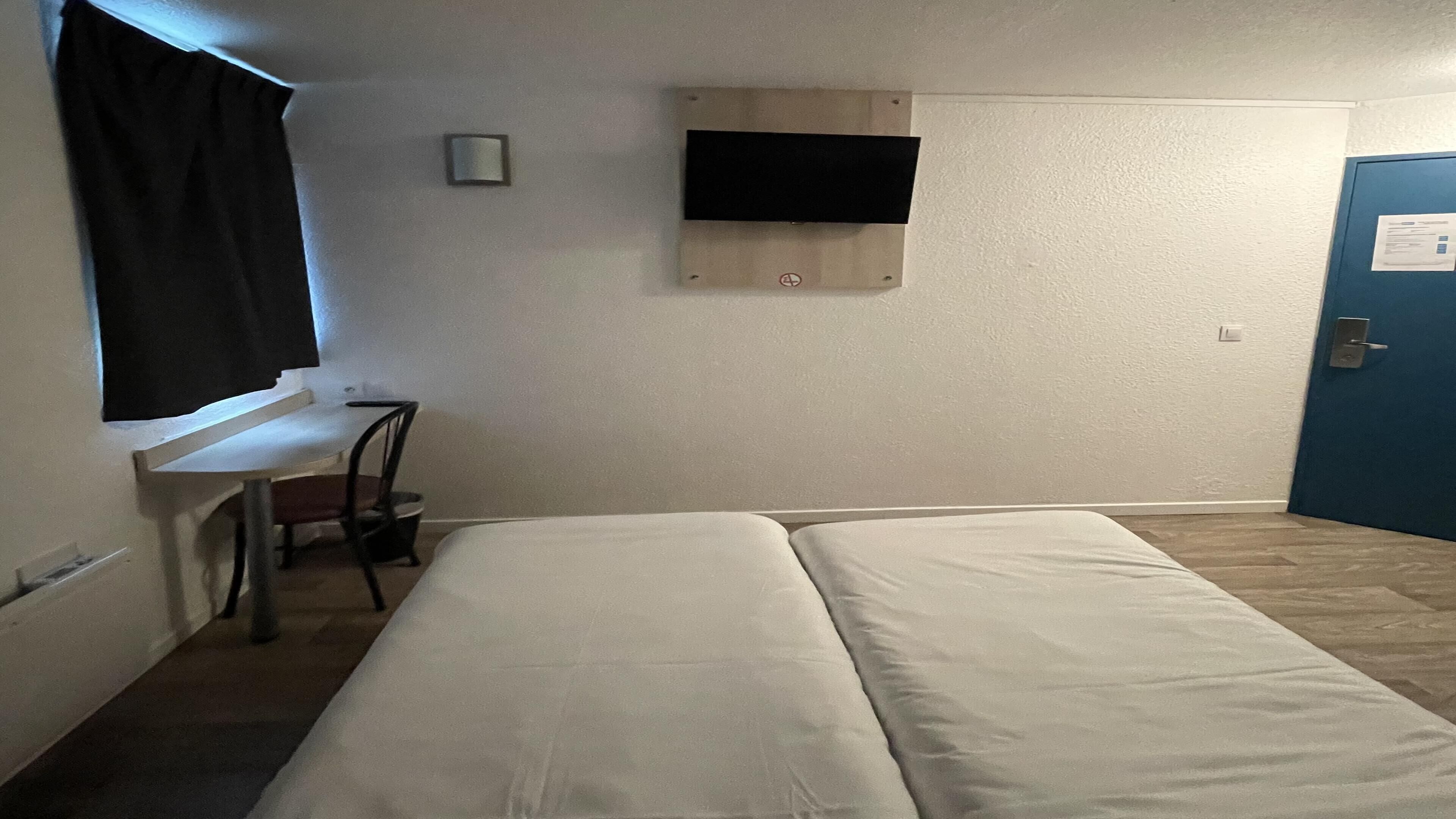Una habitación con cama, escritorio, silla, televisor montado en la pared y una puerta.