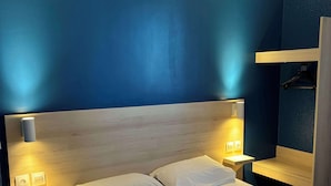 A hotel room with a large bed, wooden headboard, bedside tables with lamps, and a wooden bench. - Kyriad Direct Dreux (Dreux)