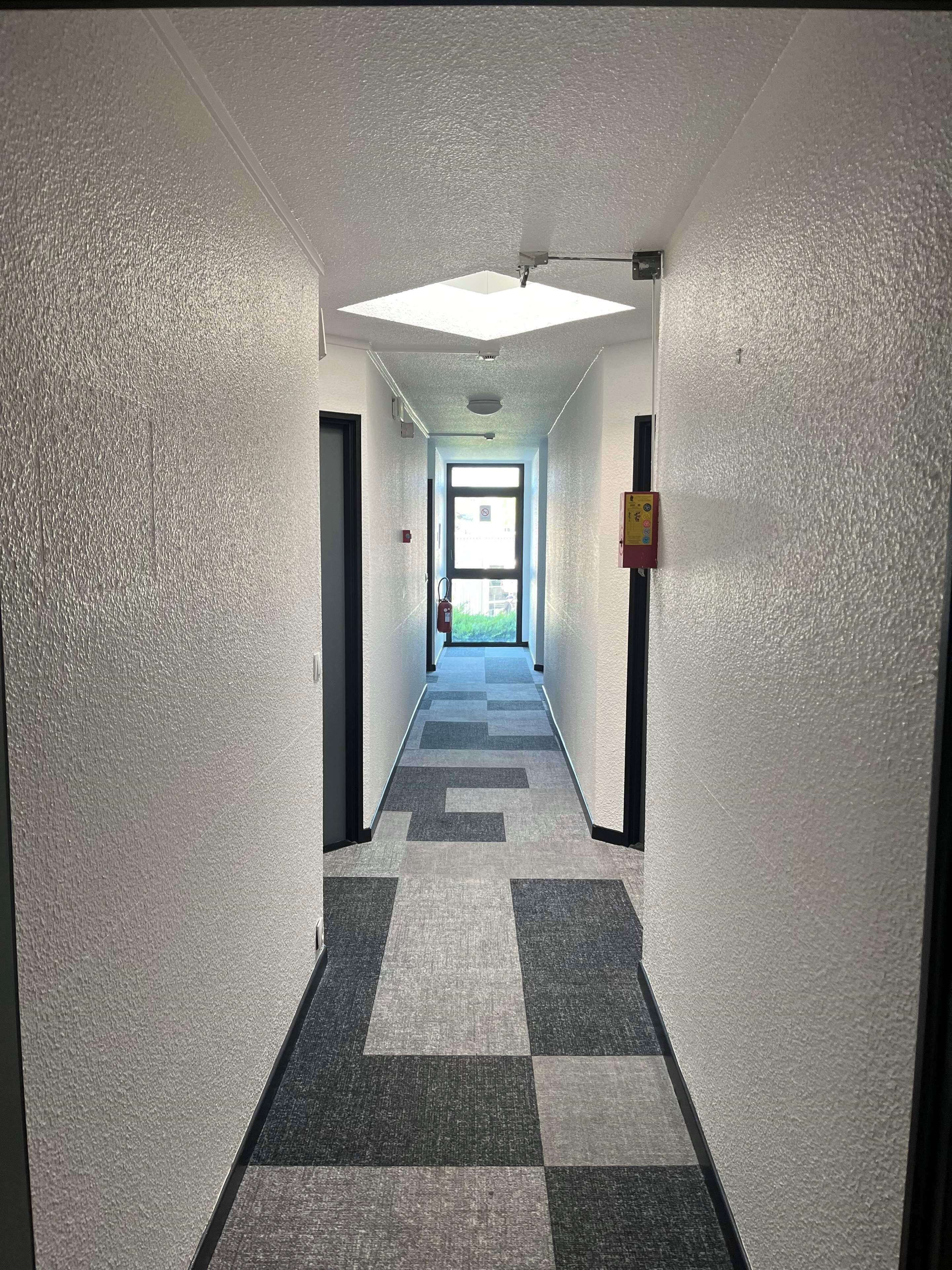 A hallway with a checkered carpet, white walls, and a door at the end leading outside.