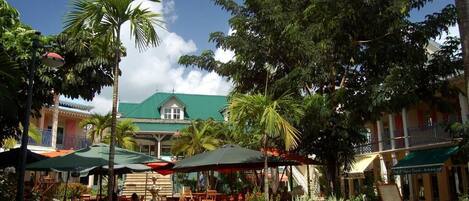 A tropical outdoor dining area with tables and umbrellas, surrounded by lush greenery and a building with a green roof.