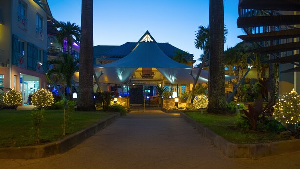 A hotel entrance with a covered walkway, palm trees, and illuminated decorations. - Village Creole (Trois-Ilets)