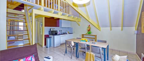 A spacious living area with a high ceiling, a dining table set with chairs, a sofa with a colorful cushion, and a small round table with a remote control.