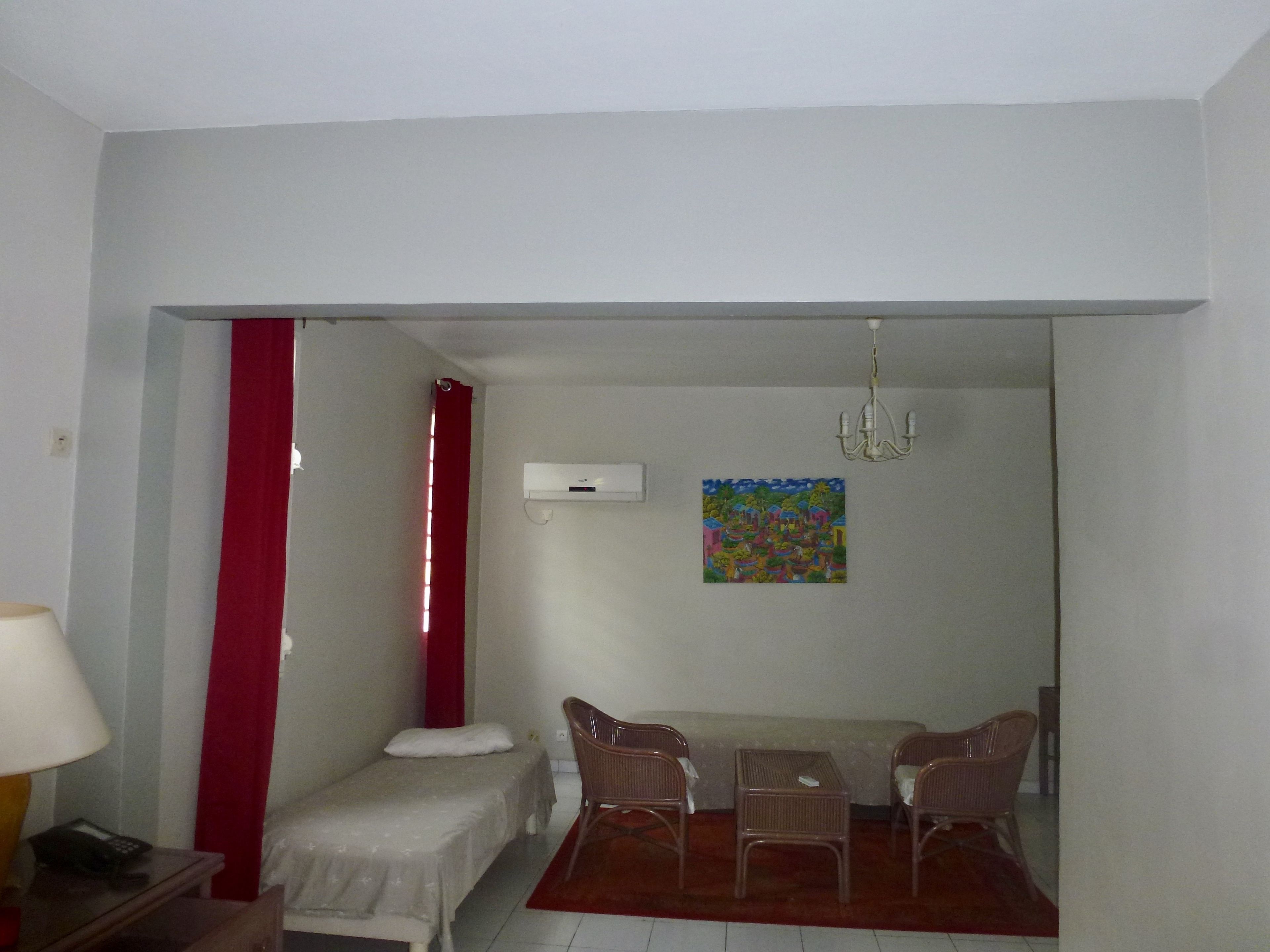 Una habitación pequeña y sencilla con cama, una mesa con sillas, una cortina roja, un cuadro en la pared y un equipo de aire acondicionado.
