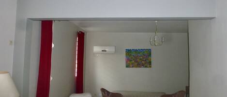Una habitación pequeña y sencilla con cama, una mesa con sillas, una cortina roja, un cuadro en la pared y un equipo de aire acondicionado.