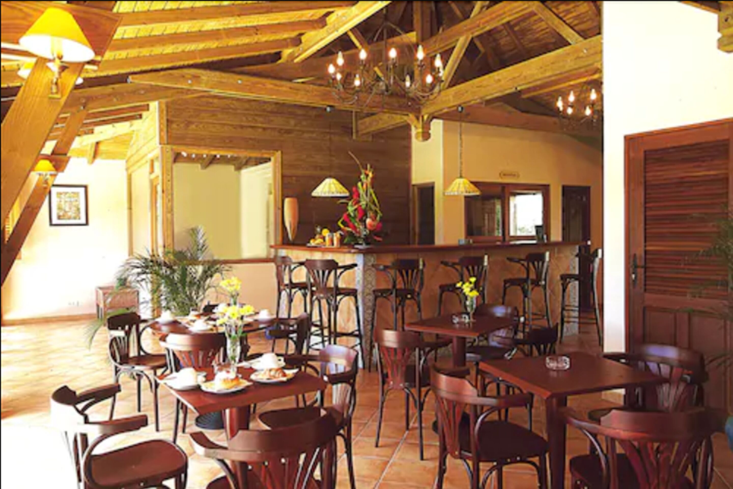 Un espace repas avec des tables et des chaises en bois, un lustre et un coin bar agrémenté d’une plante et de fleurs.