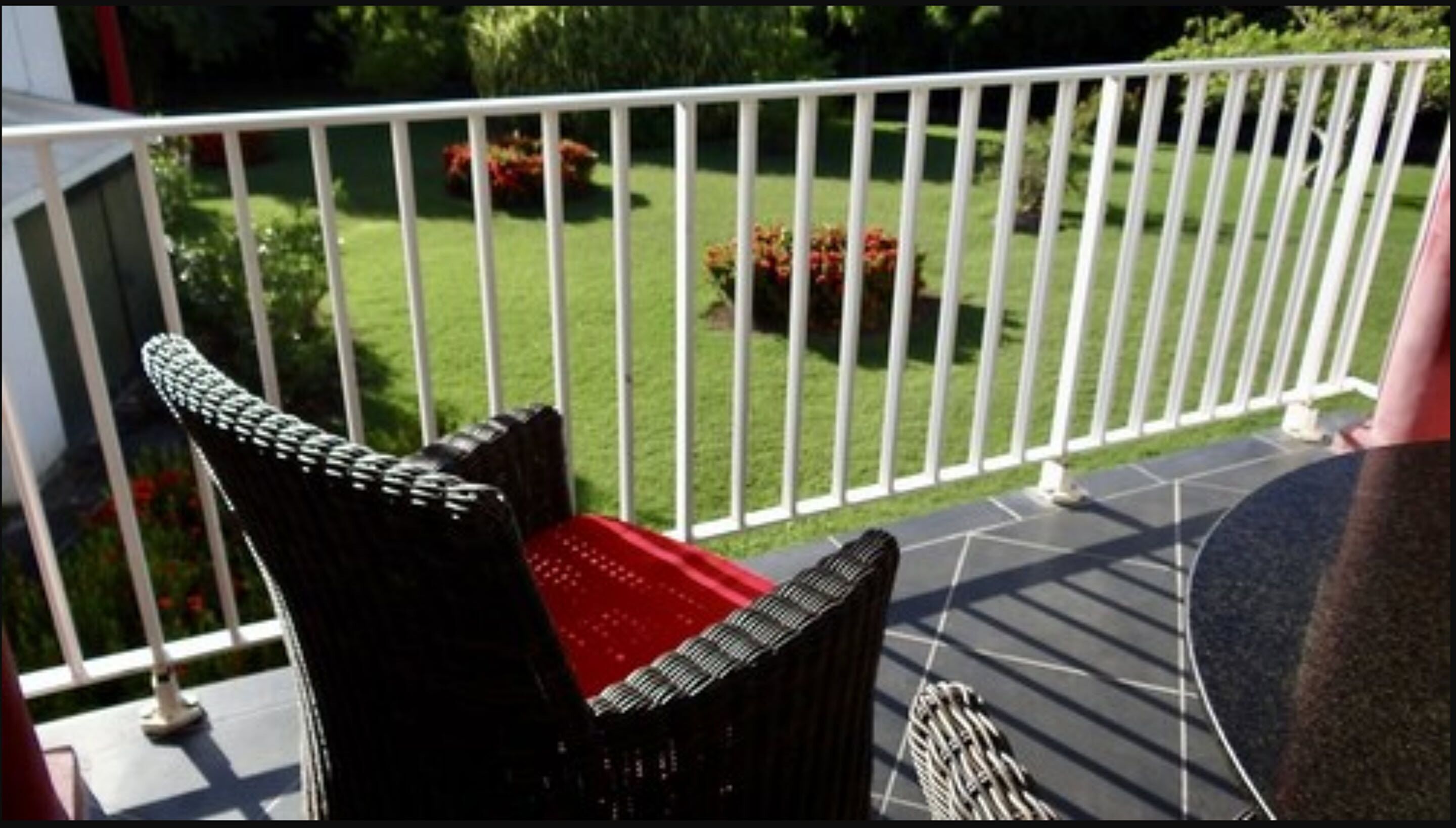 A balcony with a white railing, a wicker chair with a red cushion, and a garden with green grass and red flowers.