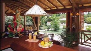 A wooden bar area with a hanging light fixture, a table with a fruit platter, and a view of the outdoors. - Résidence Le Vallon Guadeloupe (Saint-François)