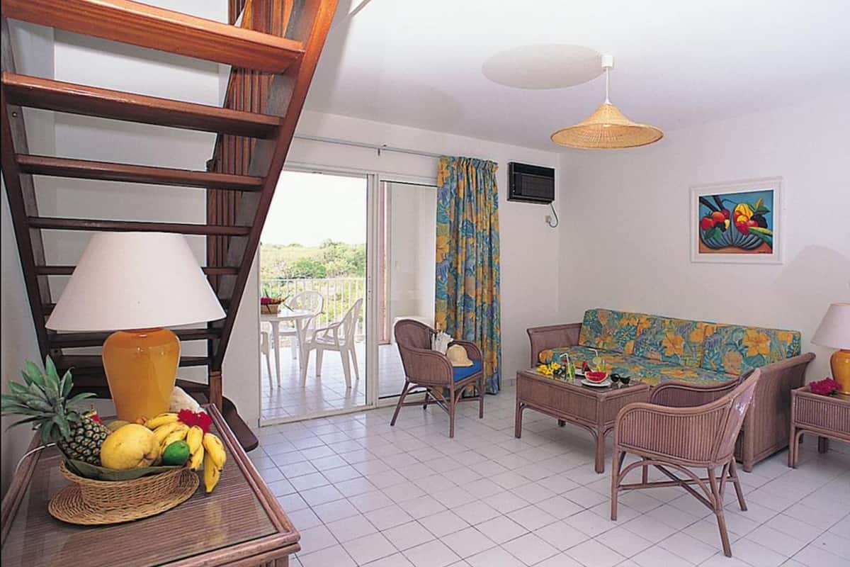 A living room with a sofa, chairs, a table with a fruit basket, a lamp, and a view of the outdoors.