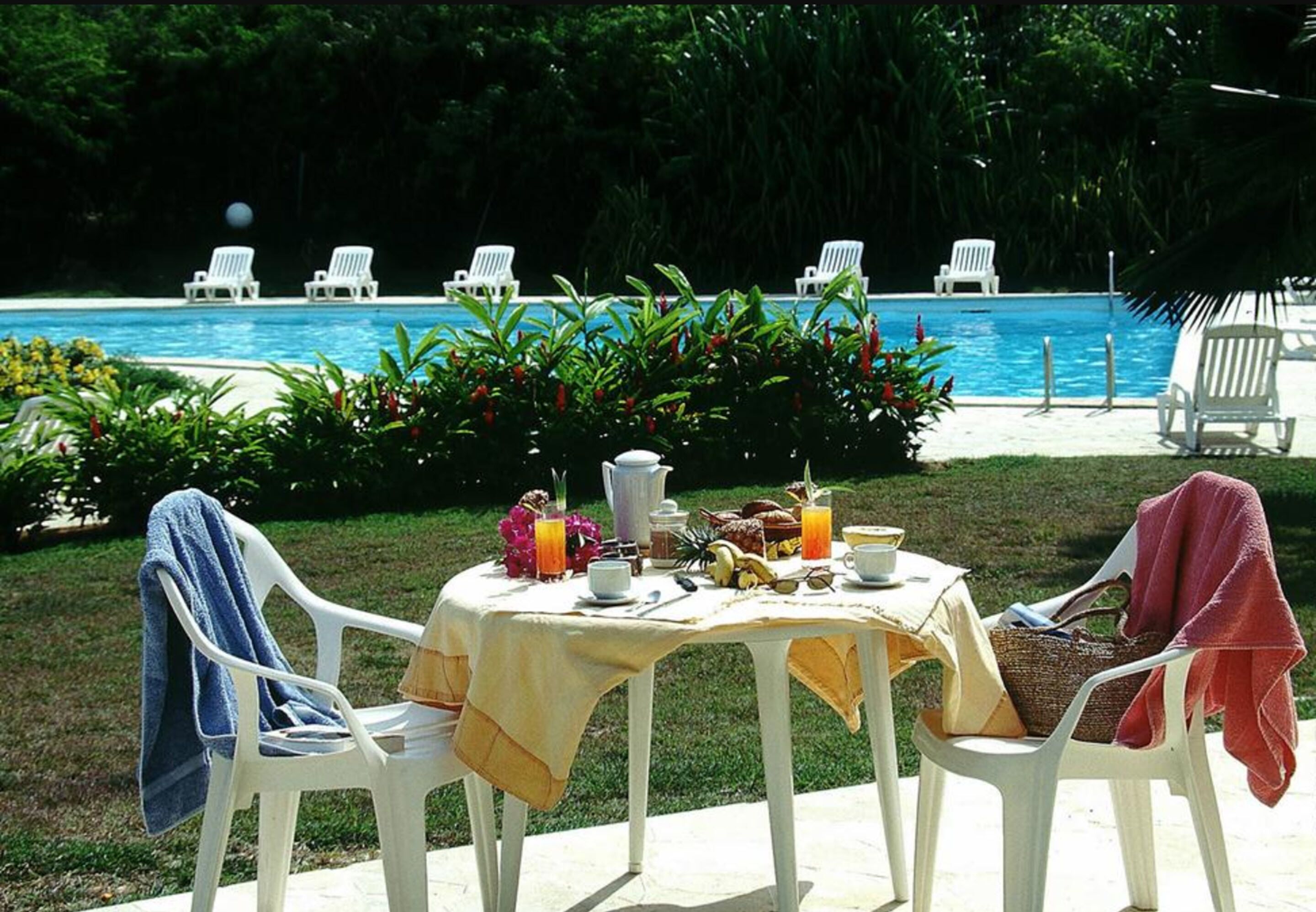 Un área de comedor junto a la piscina, con una mesa dispuesta para dos personas, rodeada de sillas blancas, cerca de una piscina.