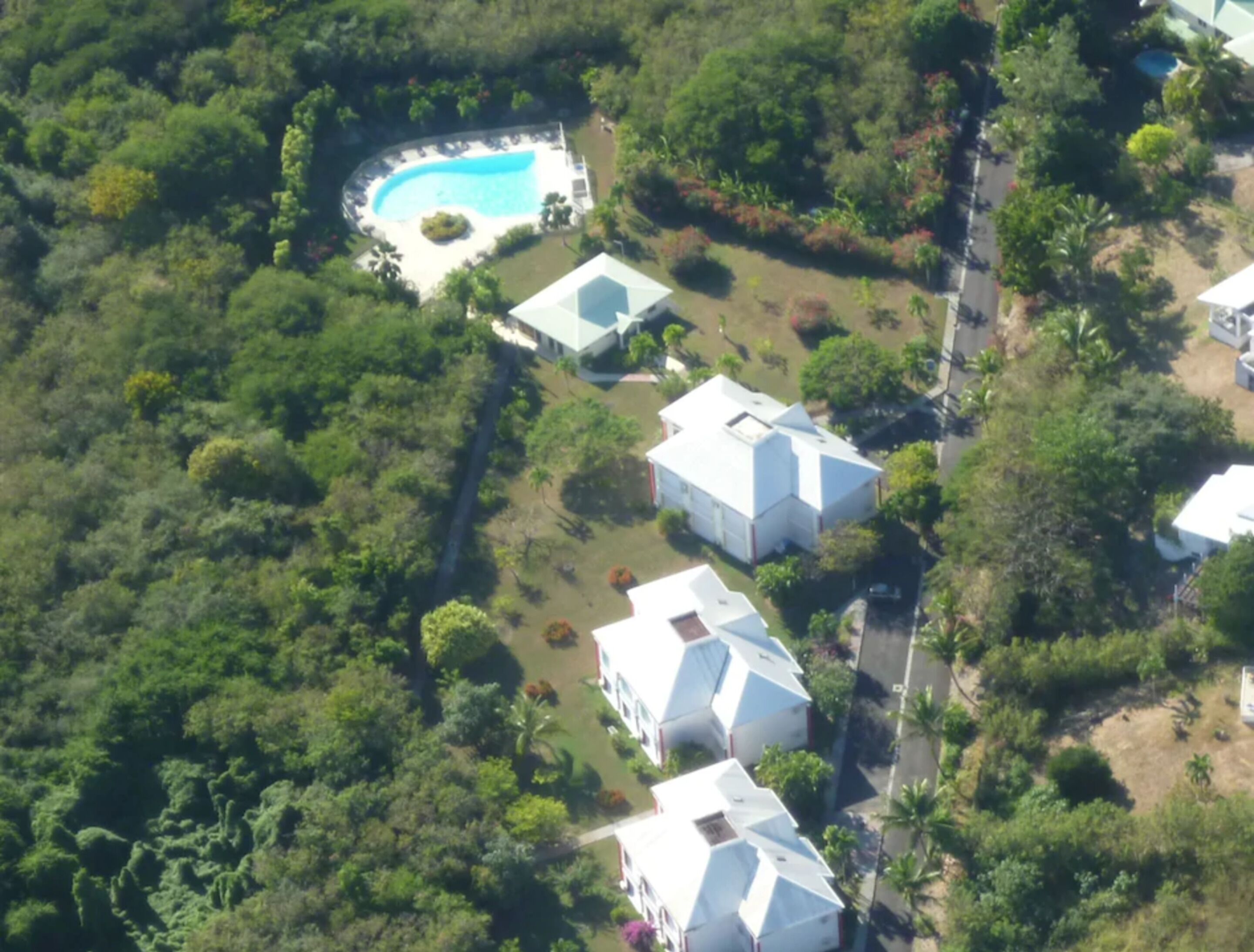 An aerial view of a residential area with a swimming pool, surrounded by lush greenery and several white houses with distinct architectural styles.