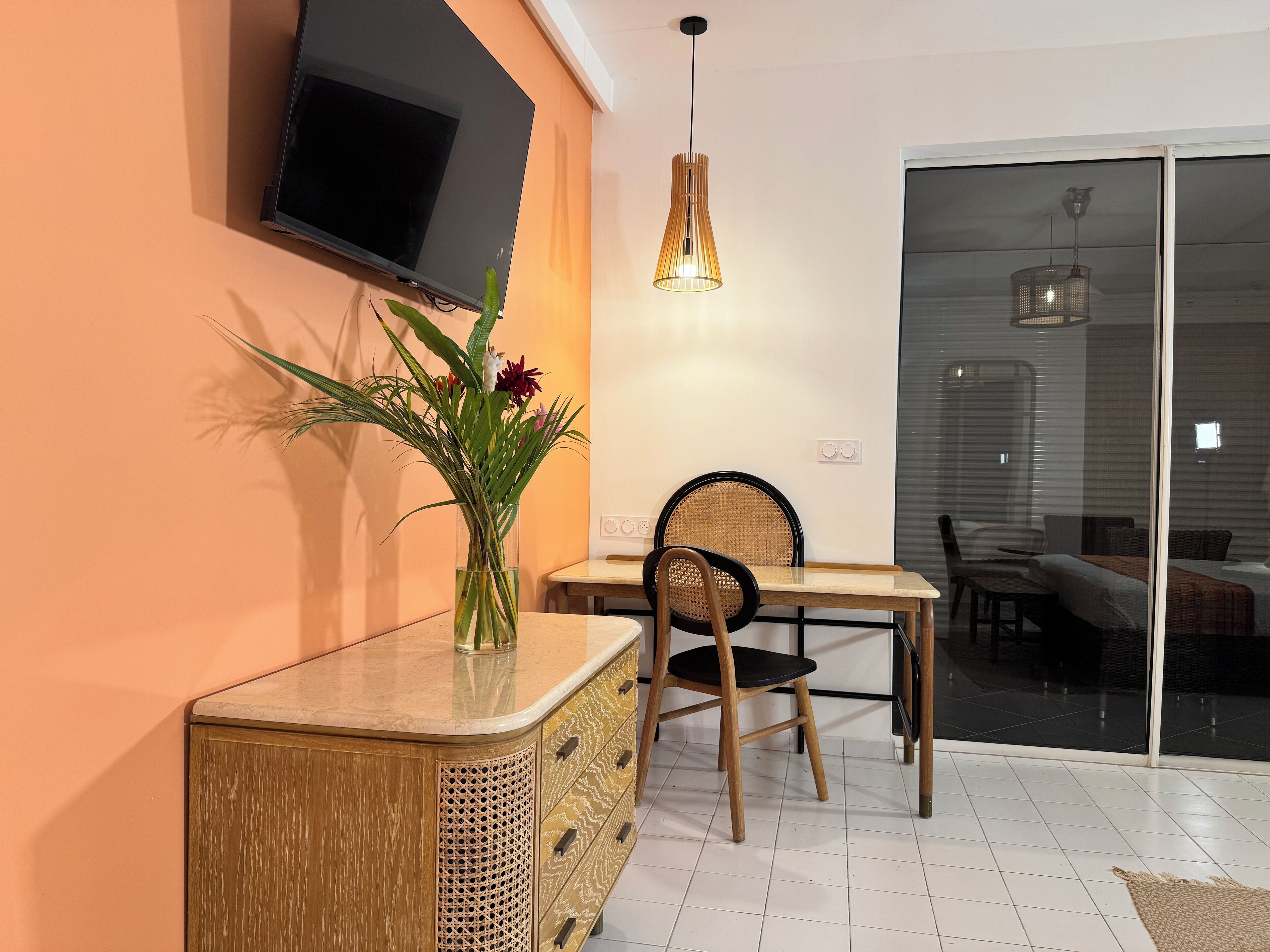 A dining area with a TV mounted on the wall, a wooden cabinet with drawers, a chair, and a table.